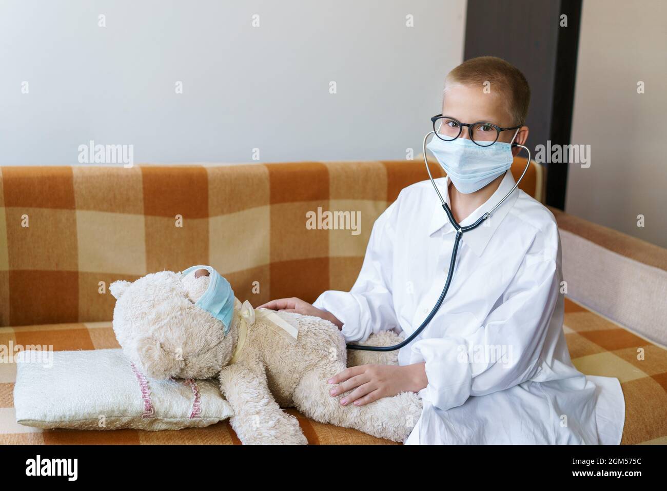 Cute little caucasian boy playing funny game like doctor holding stethoscope listening to preschooler toy pretending to be nurse treat fluffy patient at home. In a dressing gown and a medical mask. Stock Photo