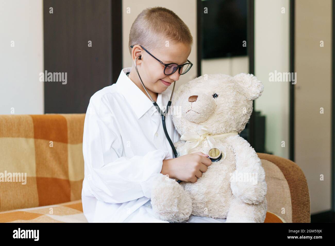 Cute little caucasian boy playing funny game like doctor holding stethoscope listening to preschooler toy pretending to be nurse treat fluffy patient at home. In a dressing gown and a medical mask. Stock Photo