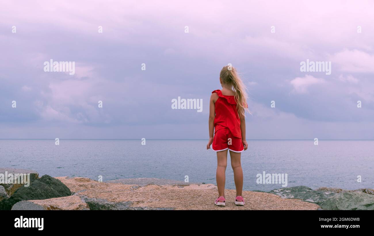 A little girl in red clothes stands with her back and looks at the endless sea and pinkish clouds on the rocky shore. Marine minimalistic landscape Stock Photo