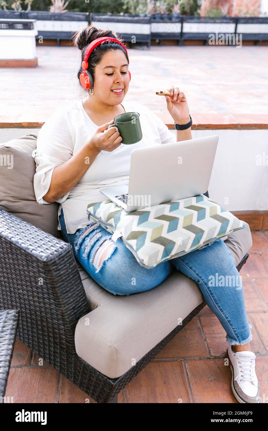 Young mexican female with headphones listening to music on computer while eating a snack on the