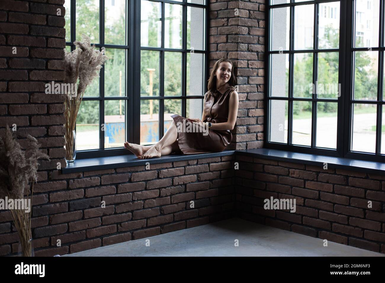 Beautiful young woman in brown leather dress sits on the windowsill ...