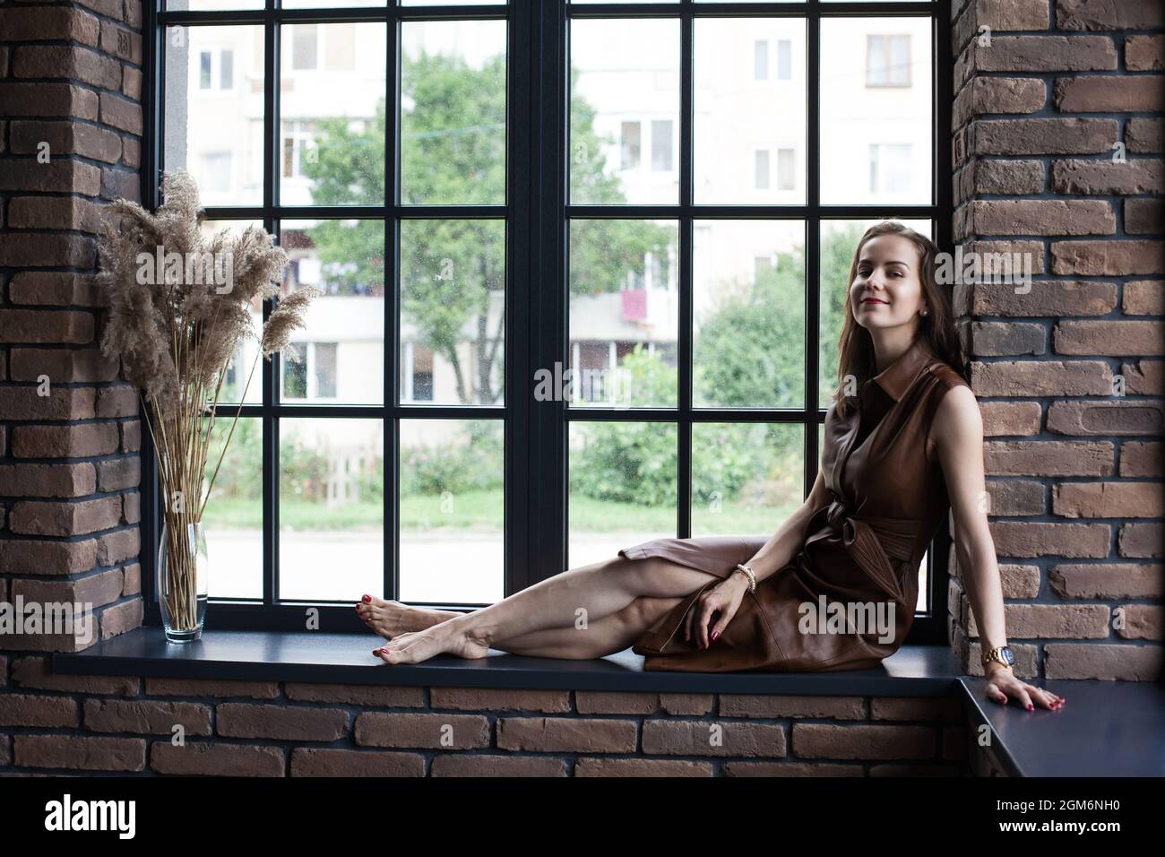 Beautiful young woman in brown leather dress sits on the windowsill ...