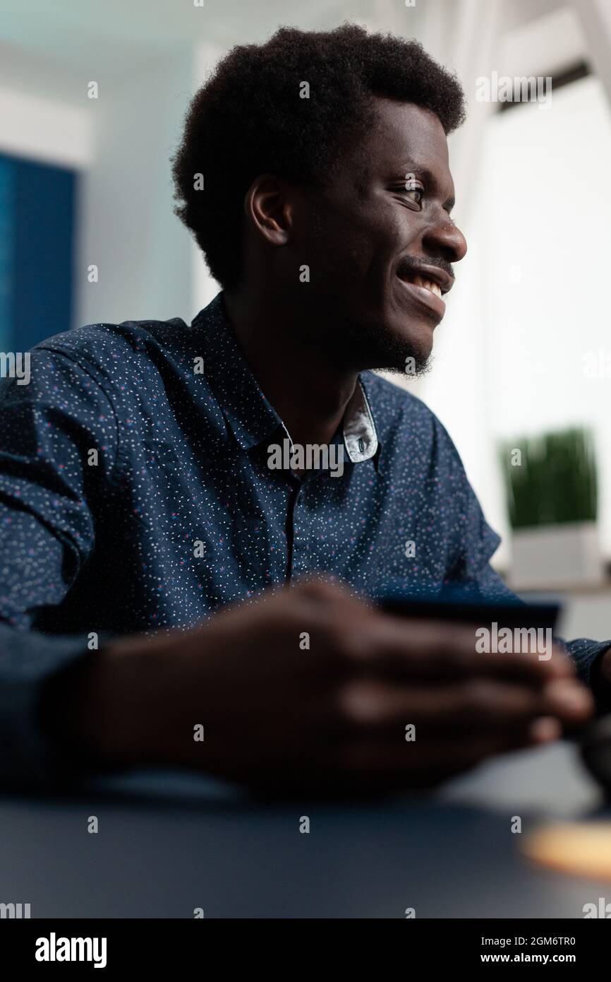 Student holding credit card paying for purchases in web store sitting at desk in living room. African american teenager doing shopping on sale website. Man paying order with electronic wallet Stock Photo