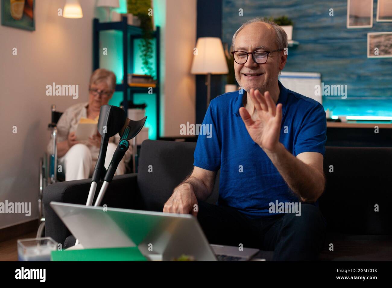 Elderly man waving at video call conference on laptop at home. Senior person using online remote communication with gadget on sofa while retired disabled woman sitting in wheelchair Stock Photo