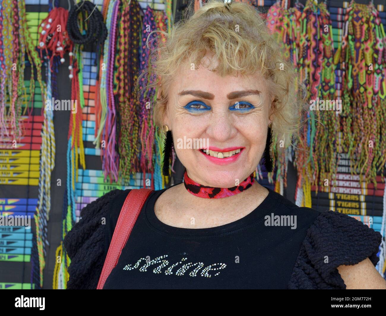 Attractive positive elderly Caucasian woman with heavy eye makeup and blond dyed hair smiles and poses in front of a colorful souvenir display. Stock Photo