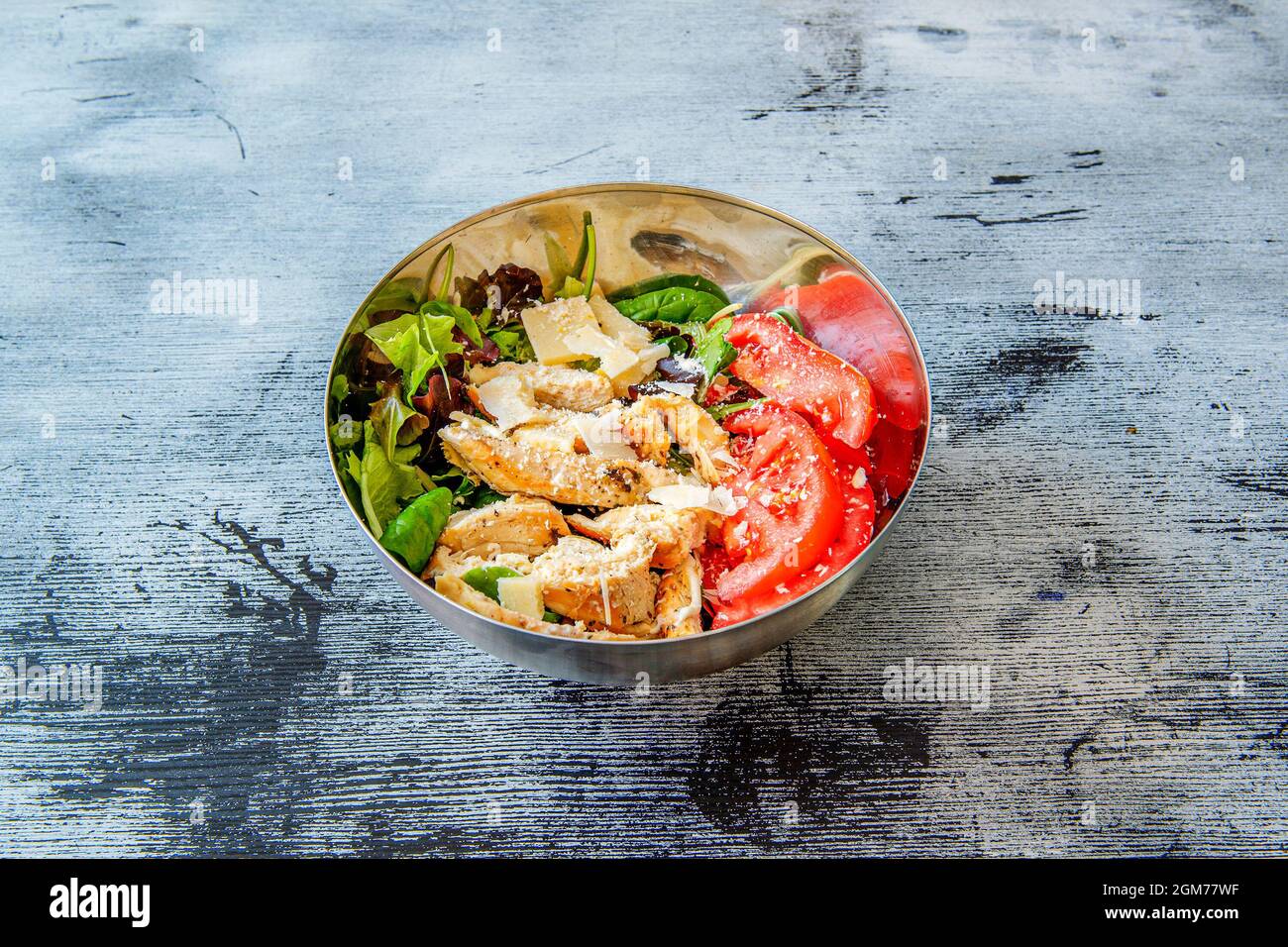 metal bowl with roast chicken, parmesan cheese flakes, special sliced tomatoes, leaves of lettuce sprouts and white rice at the base on a blue and bla Stock Photo