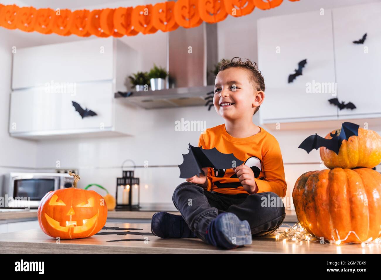 Cute child sitting at the kitchen table dressed as a pumpkin playing with bats and decorated halloween pumpkin Stock Photo