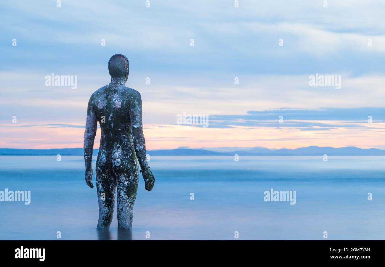 An Iron Man figure surrounded by water as the tide comes in at Another Place (near Crosby, Liverpool) at twilight in September 2021. Stock Photo