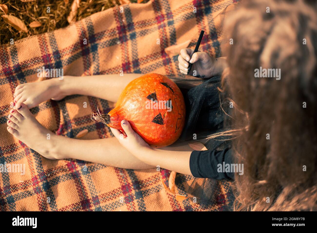 Halloween. Girl with long hair wearing black dress sits on an orange blanket in the park and draws a face on a pumpkin. Stock Photo