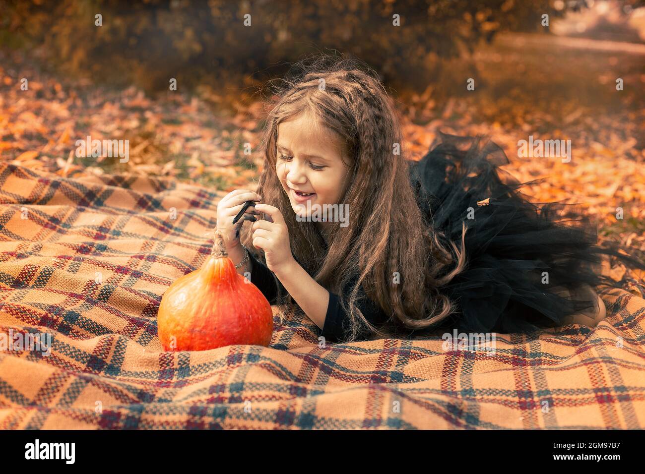 Halloween. Girl with long hair wearing black dress sits on an orange blanket in the park and draws a face on a pumpkin. Stock Photo