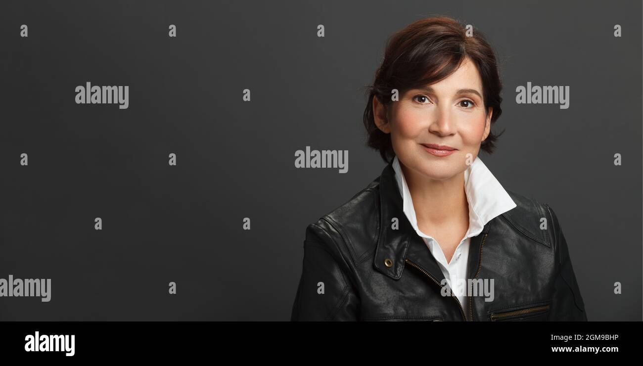 Middle aged well looking woman portrait. Woman wearing white shirt and black leather jacket against dark solid background. Studio female portrait. Bil Stock Photo
