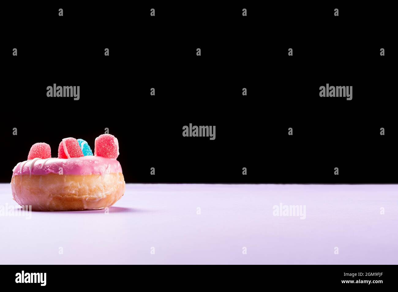 Photograph of a pink donuts decorated with jelly beans and drawn with white chocolate.The photo is taken in horizontal format on a lilac cardboard and Stock Photo