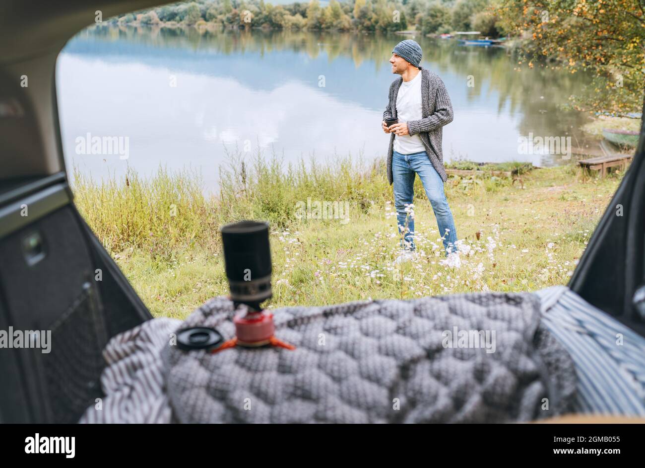 A man dressed warm knitted clothes enjoying gas stove prepared coffee and beautiful autumnal mountain lake view. Opened Car trunk shot. Warm early aut Stock Photo
