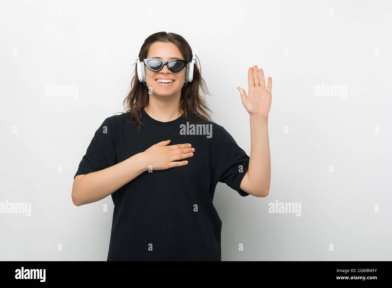 Young woman is making the promise or swear gesture while wearing headphones and sunglasses over white background. Stock Photo