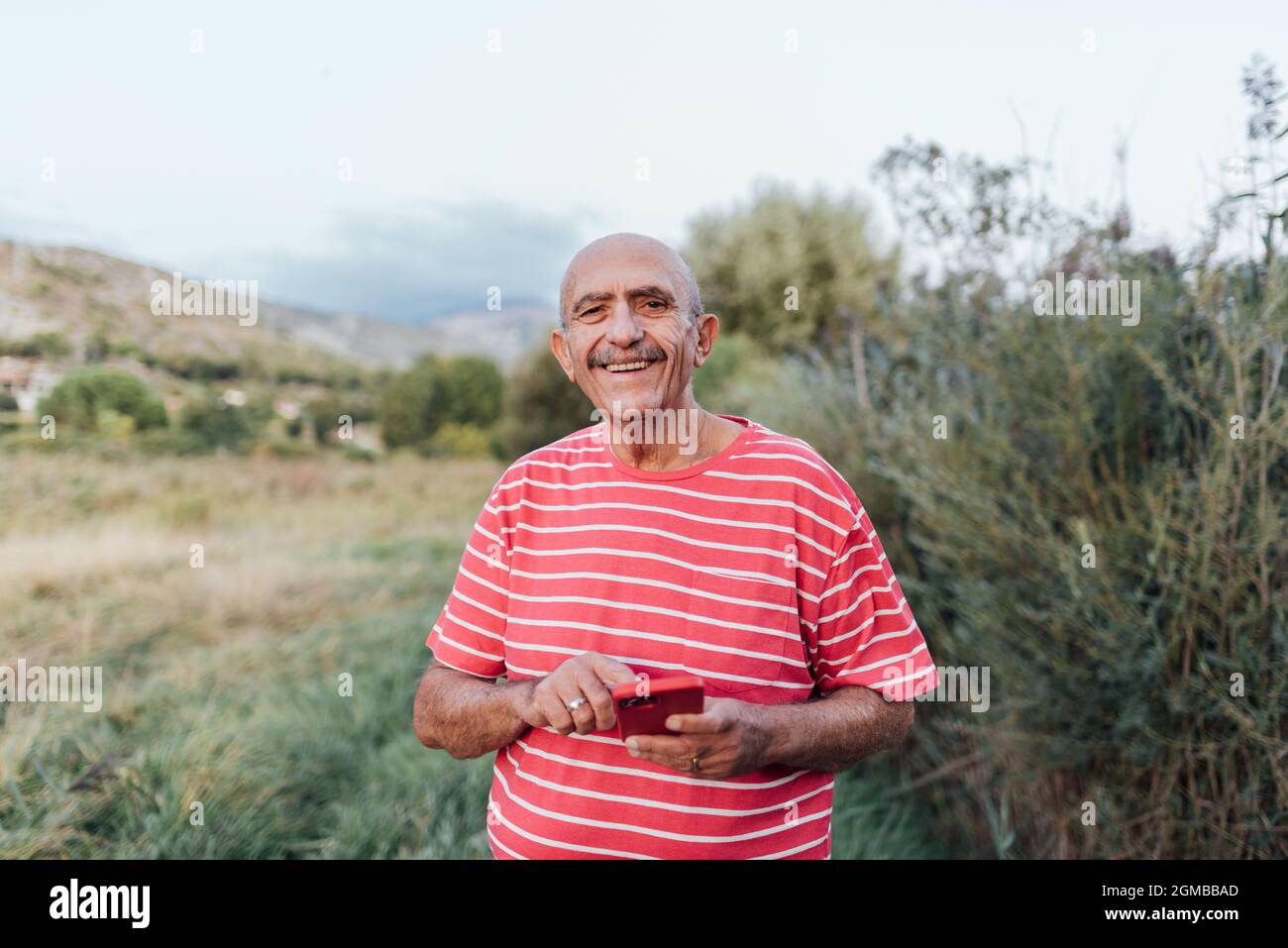 Middle-aged man looking at the camera and smiling while using a mobile phone outdoors in the countryside. Stock Photo