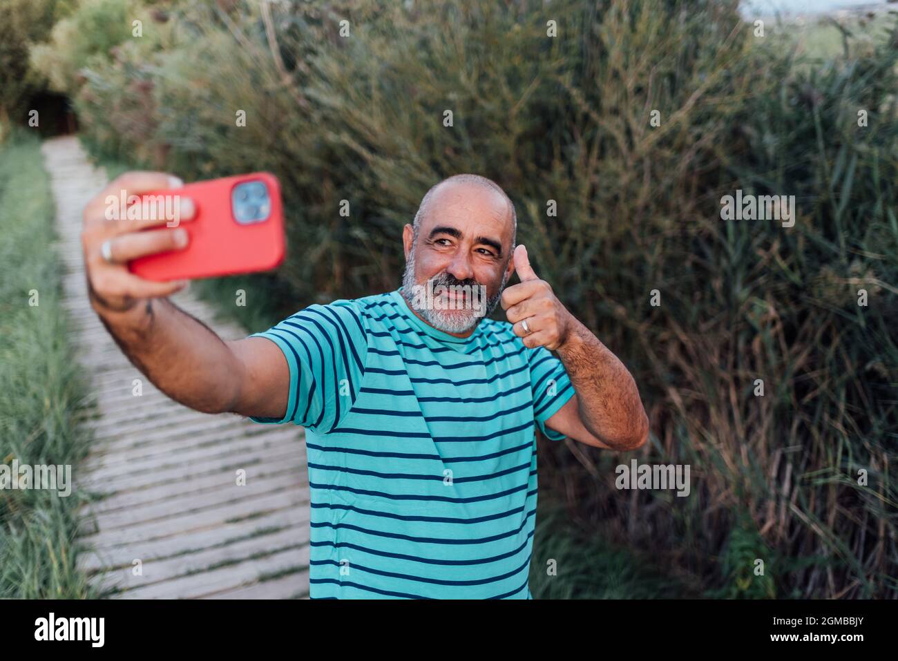 Happy middle-aged man showing thumb up while taking a selfie with a mobile phone outdoors in the nature. Stock Photo