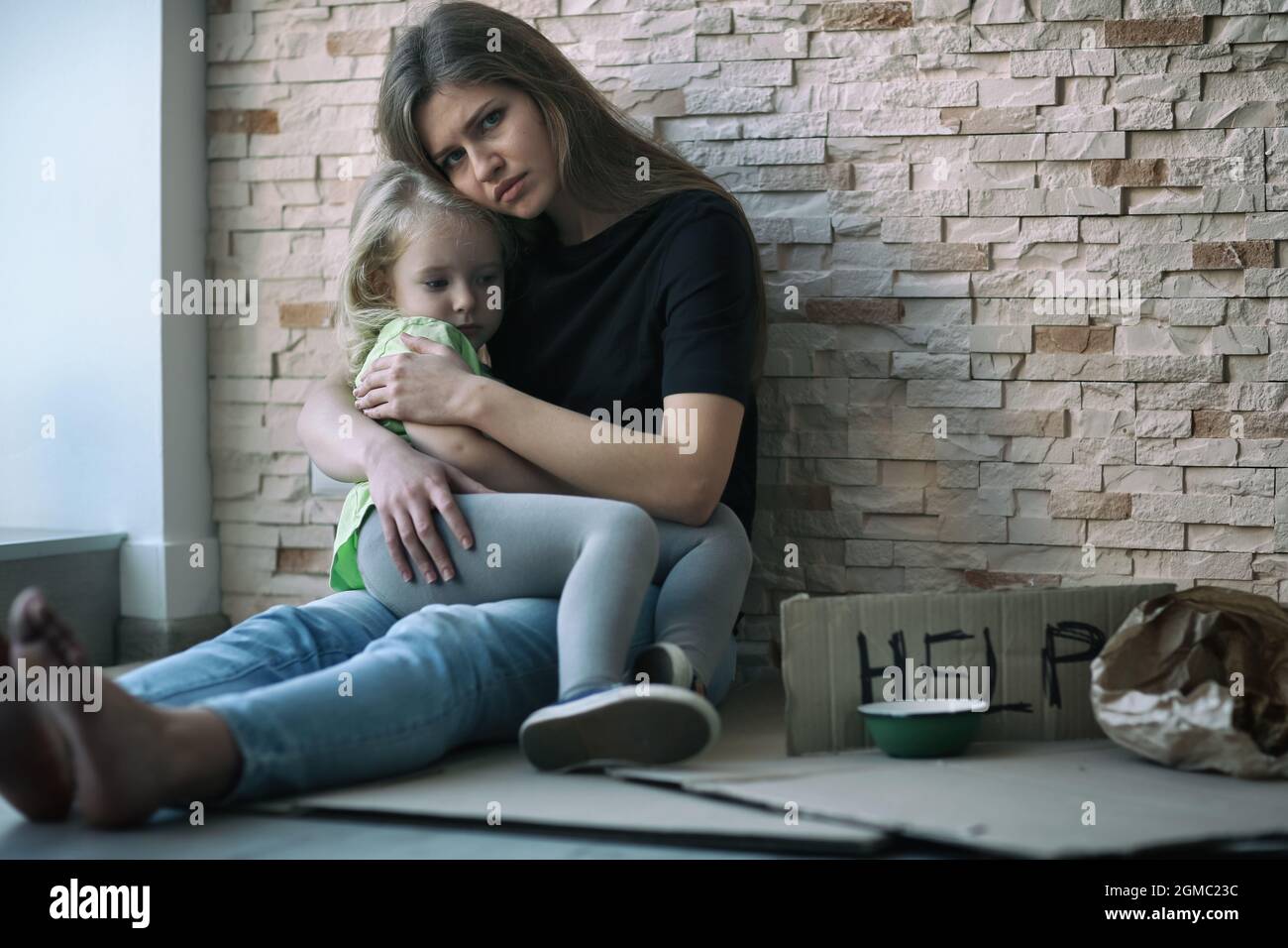 Homeless poor woman and her little daughter sitting near brick wall and