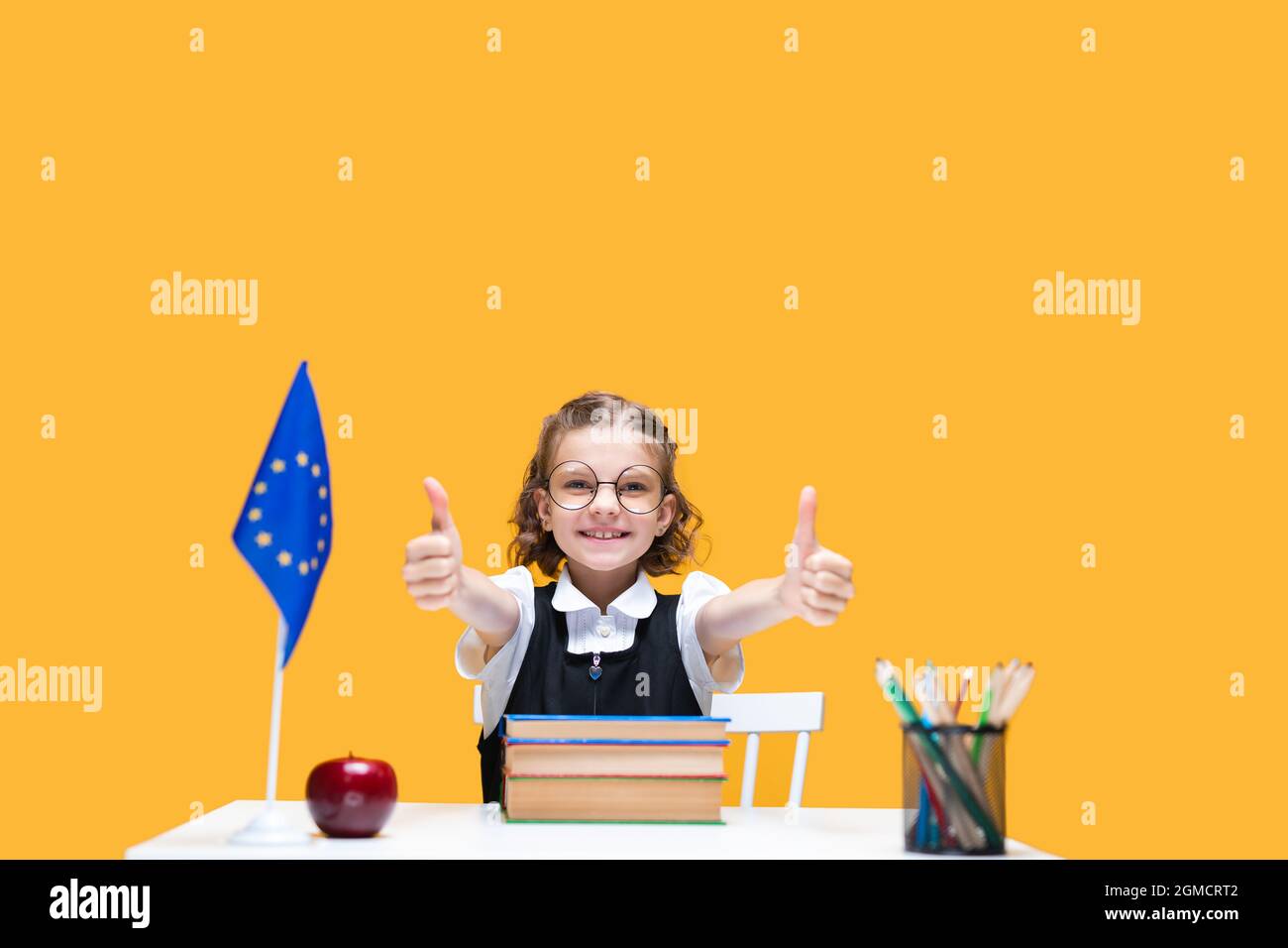Happy caucasian schoolgirl sitting at the desk gesturing thumbs up ...