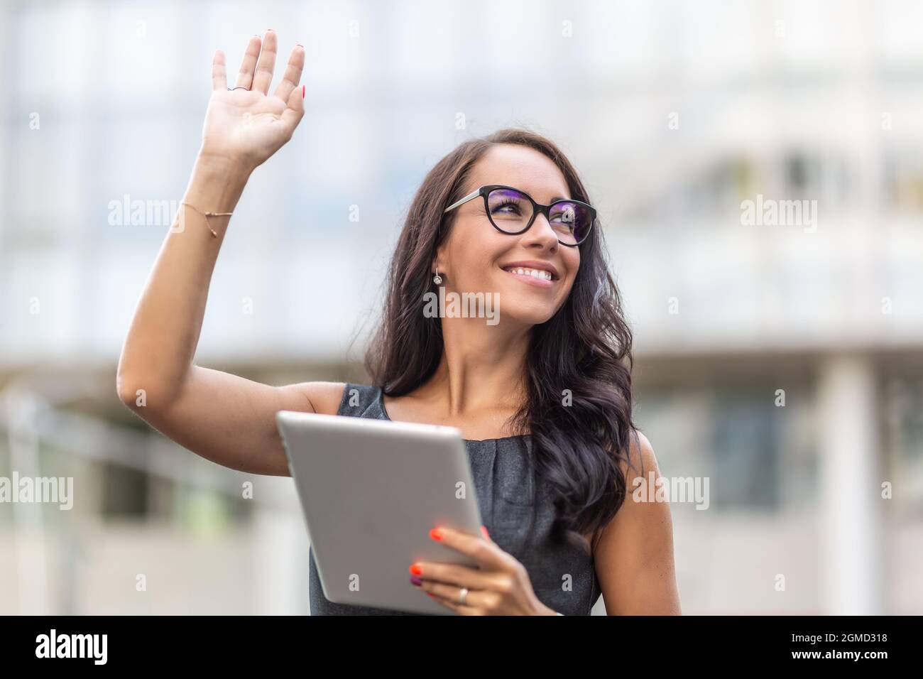 Woman in office clothes and glasses smiles and waves holding a tablet. Stock Photo