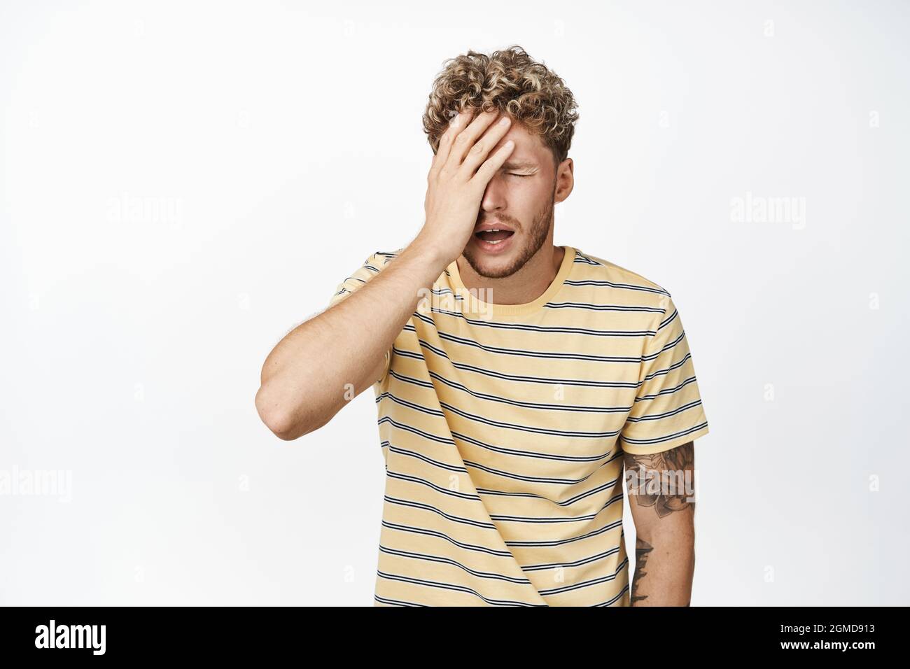 Shocked distressed blond man making facepalm, standing frustrated and upset, crying and sobbing, standing in casual clothes over white background Stock Photo