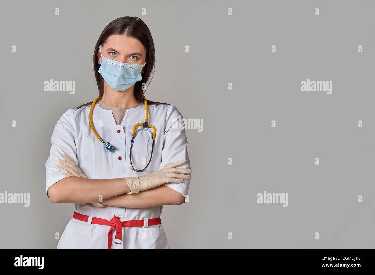 A young female doctor in a white coat with a stethoscope and a medical mask on a gray isolated background. copy space Stock Photo