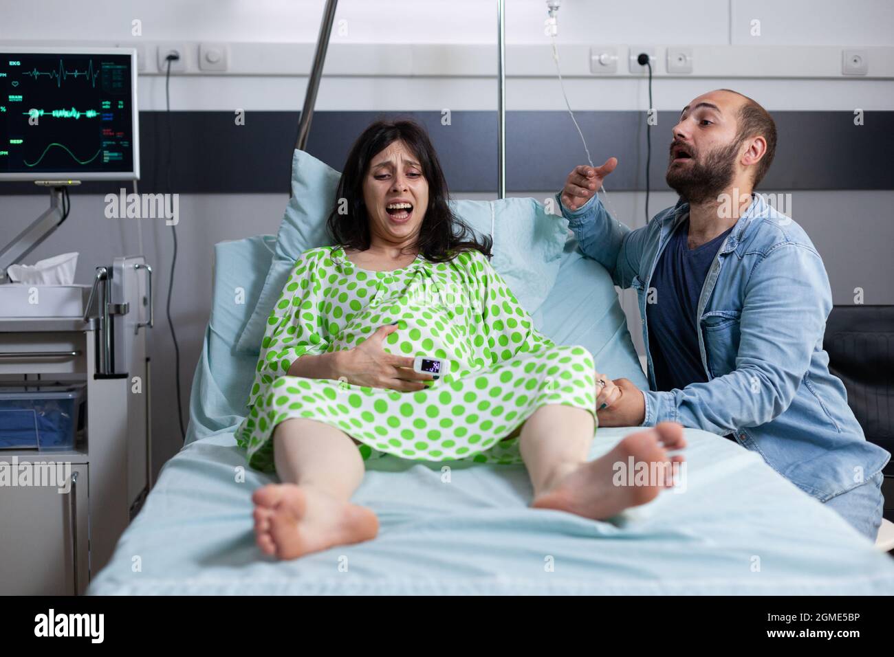 Caucasian couple expecting baby in hospital ward bed at medical facility. Pregnant woman with painful contractions getting into labor while young man panicking about childbirth. Stock Photo