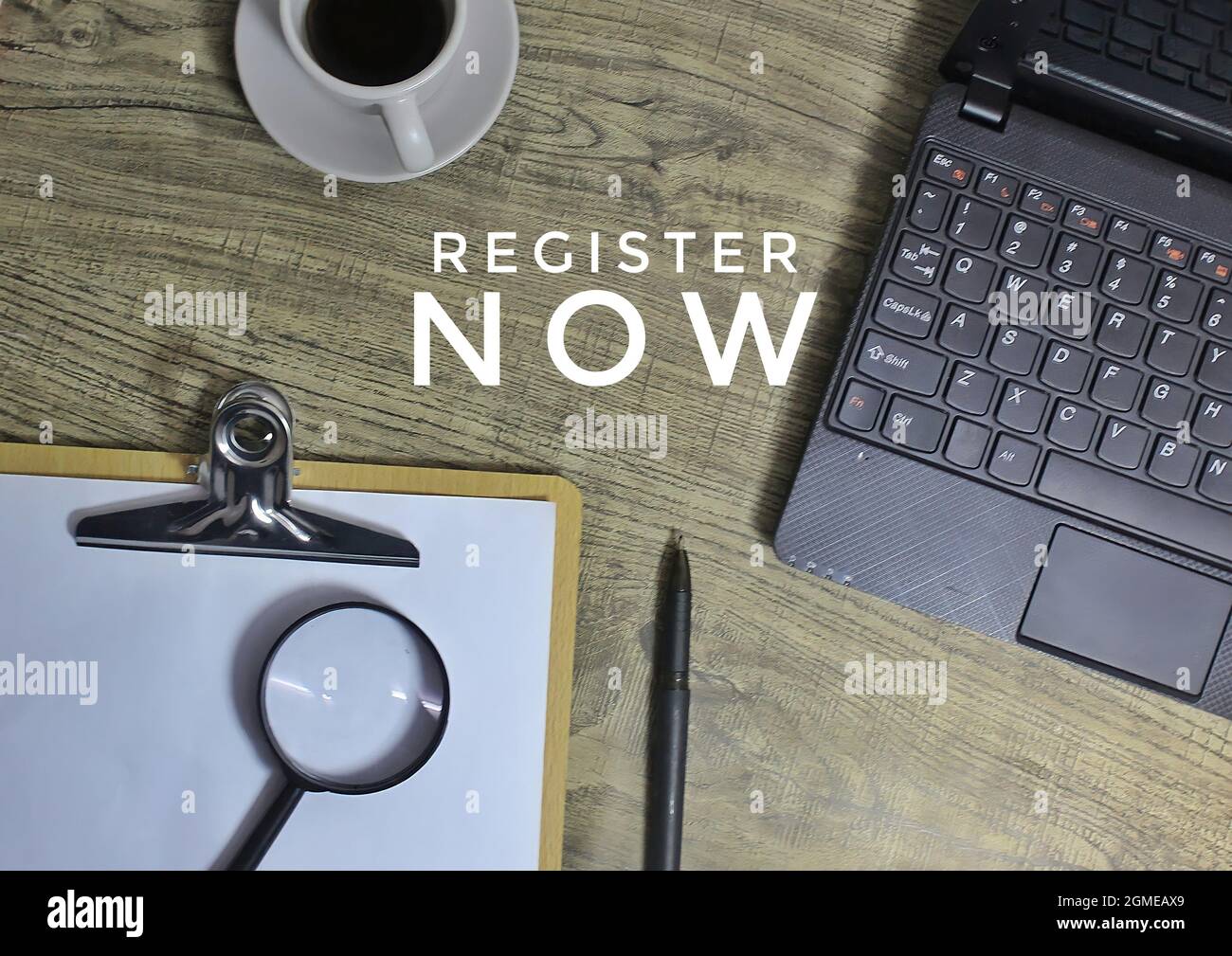 Top view of text REGISTER NOW, coffee, laptop, pen and magnifying glass. Stock Photo