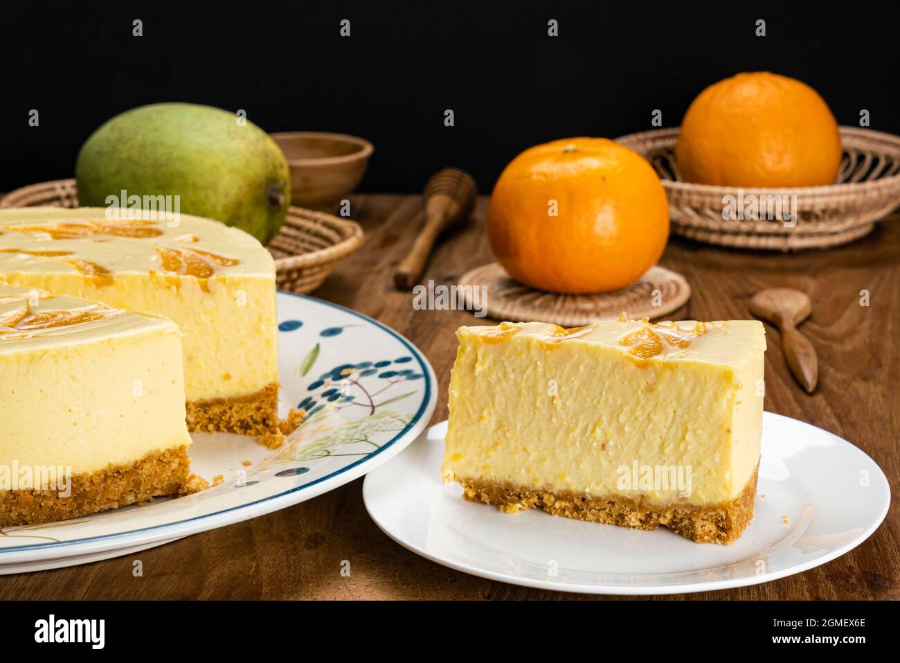 Putting a portion of delicious homemade mango cheesecake in white ceramic dish with a big loaf of cake in ceramic dish, mango, ripe oranges, wooden sp Stock Photo