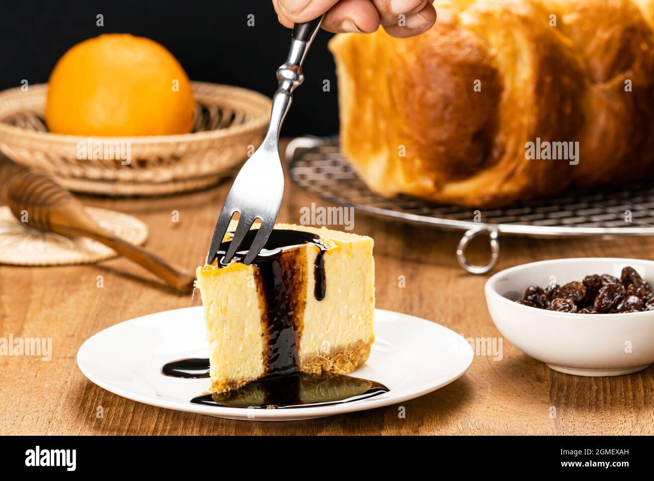 Using metal fork taking a bite of delicious homemade mango cheesecake topping with chocolate in white ceramic dish with honey dipper, loaf of bread on Stock Photo