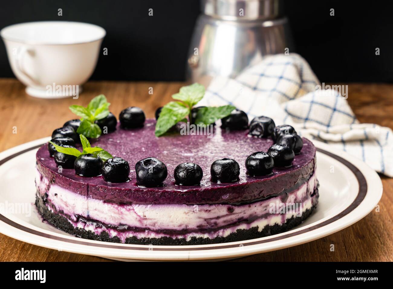 Homemade blueberry cheesecake garnished with preserved blueberry and mint leaf in brown ceramic dish with metal moka pot coffee maker and white coffee Stock Photo