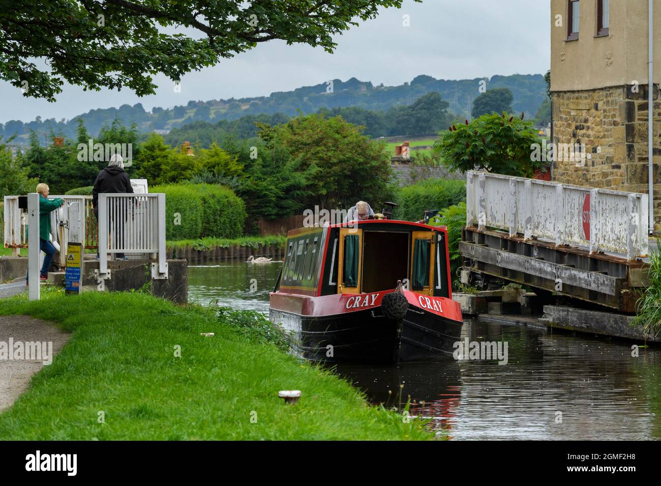 People watch hire narrowboat travelling past & through Micklethwaite swingbridge, manual barriers open - Leeds Liverpool Canal, Yorkshire, England UK. Stock Photo