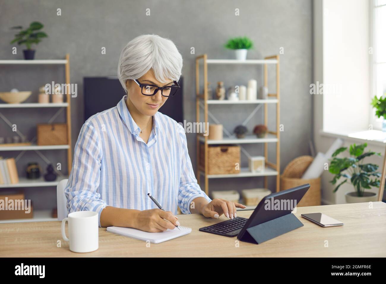 Mature woman sitting at her desk at home, using tablet and taking notes in notebook Stock Photo