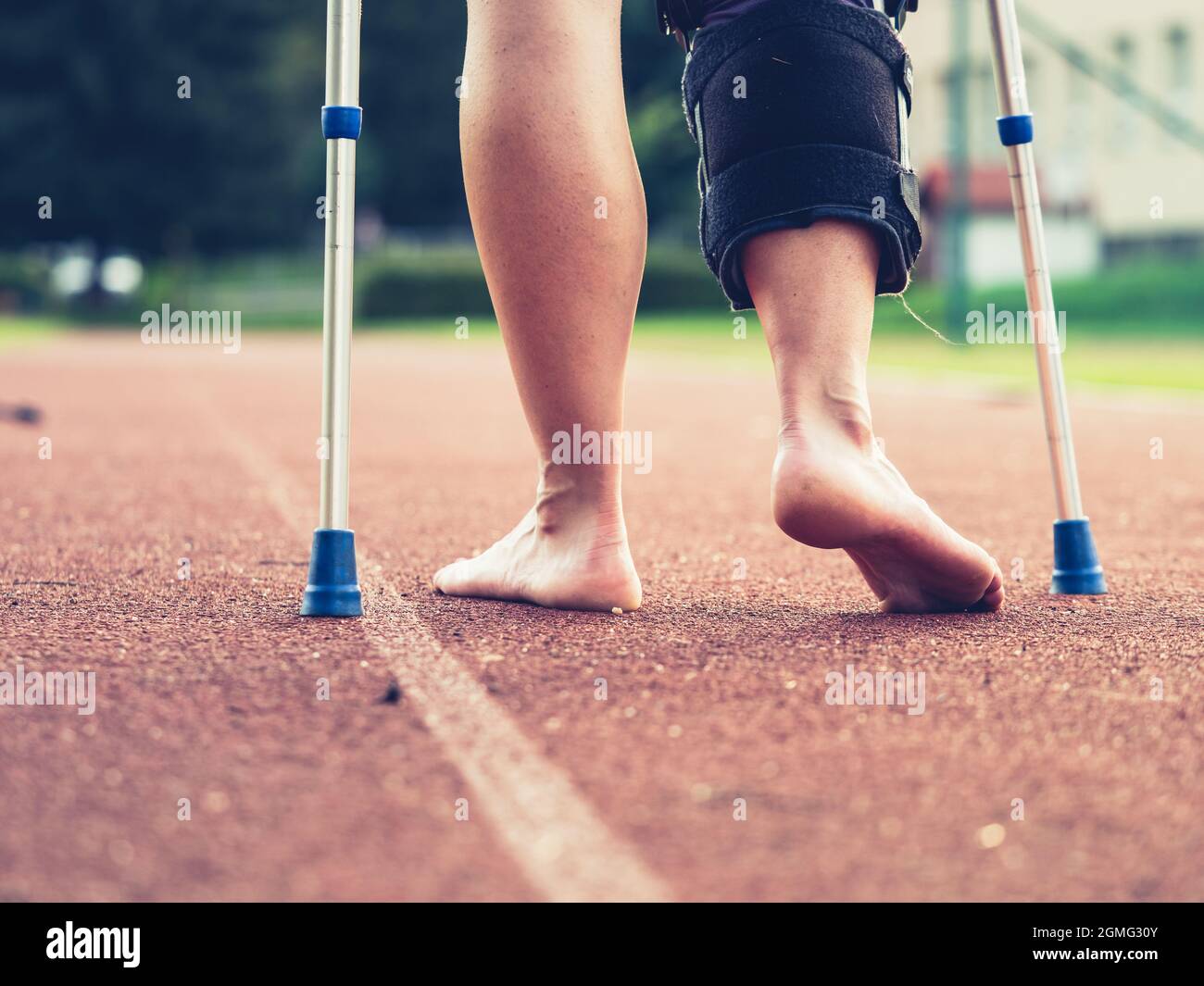Woman athlete on crutches, wearing a wrist brace and knee support, bandaged leg.  Woman walks on red running surface Stock Photo