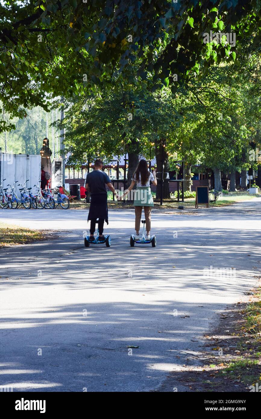 A guy and a girl ride on hoverboards in a summer park with their backs and hold hands Stock Photo