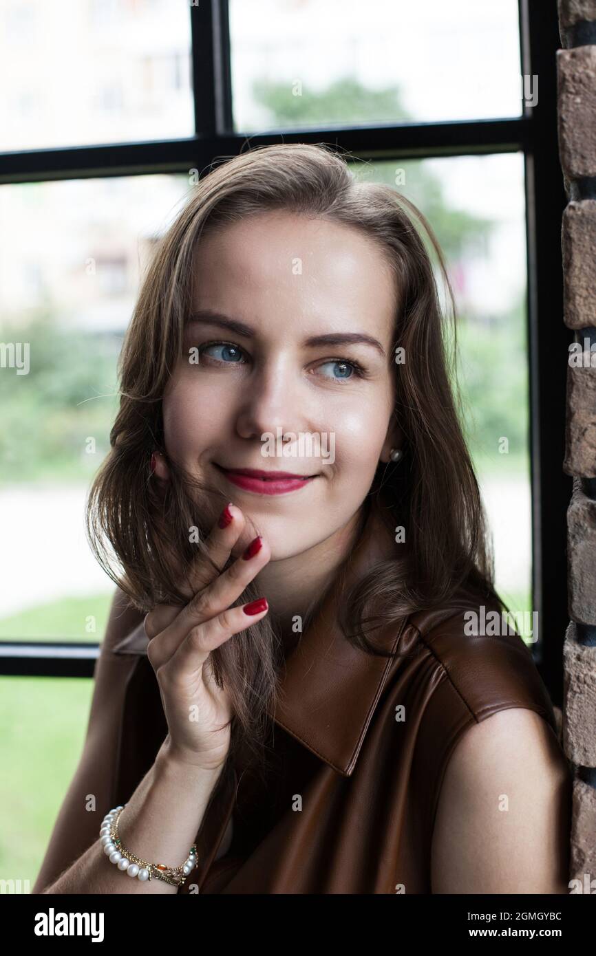 Beautiful young woman in brown leather dress sits on the windowsill ...