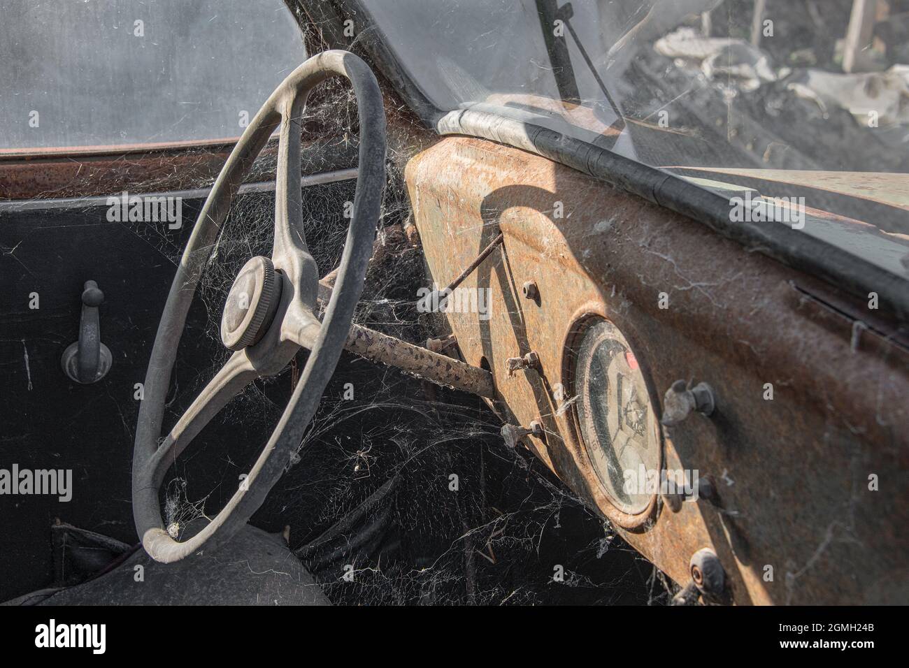 a close up image of an old retro rusty car. It is a left hand drive and shows the steering wheel and dash covered in rust and cobwebs Stock Photo