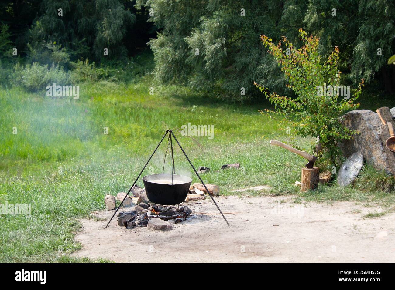 Old cast-iron cauldron on a fire. Cooking food outdoors in a pot on a tripod. Ax in a wooden stump. Medieval Knight Shield and copper ladle Stock Photo