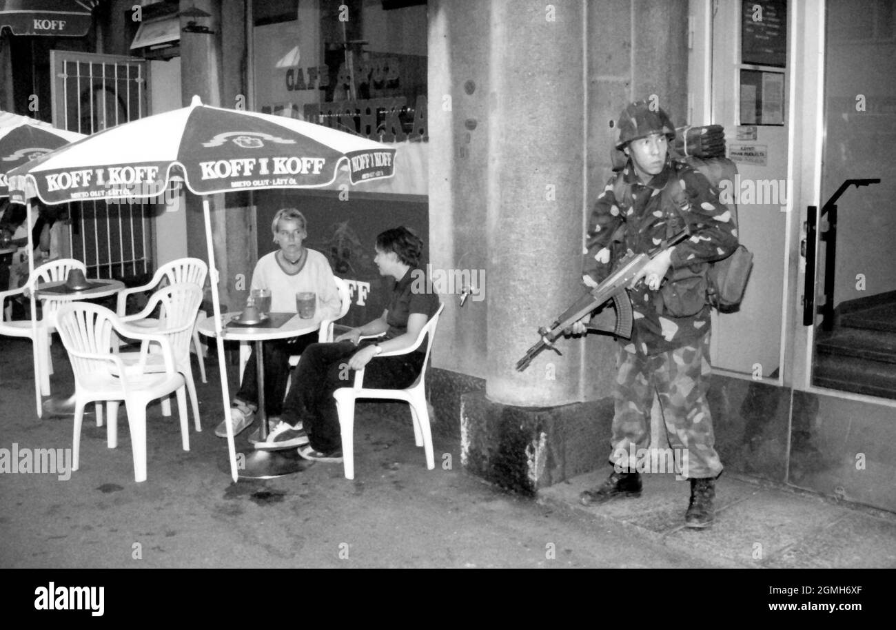 Infantry soldier, armed with assault rifle, on a sidewalk during a military exercise in Hakaniemi, Helsinki, Finland, circa 1996. Adjacently, two people having a drink at a bar table under a parasol. Stock Photo