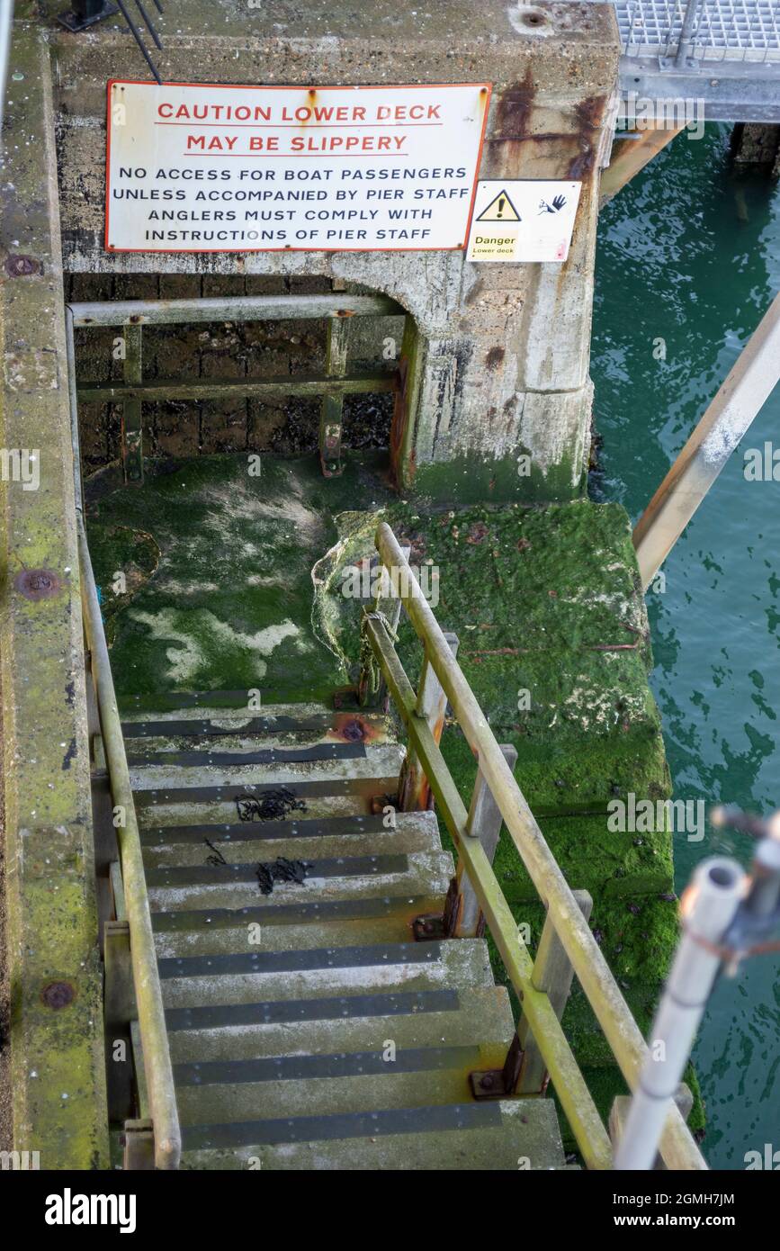 Disused steps down to the sea on Southend Pier once used to board a boat or vessel. Warning sign. Caution, lower deck may be slipper. Algae covered Stock Photo