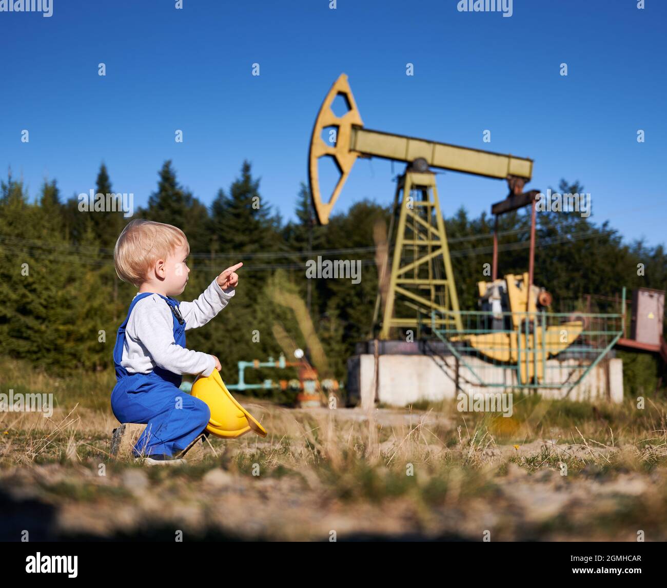 Side view of child boy pointing finger up to petroleum pump jack. Young future oilman examining for oil extraction from subsoil of the earth by rocking pump in petroleum field. Stock Photo