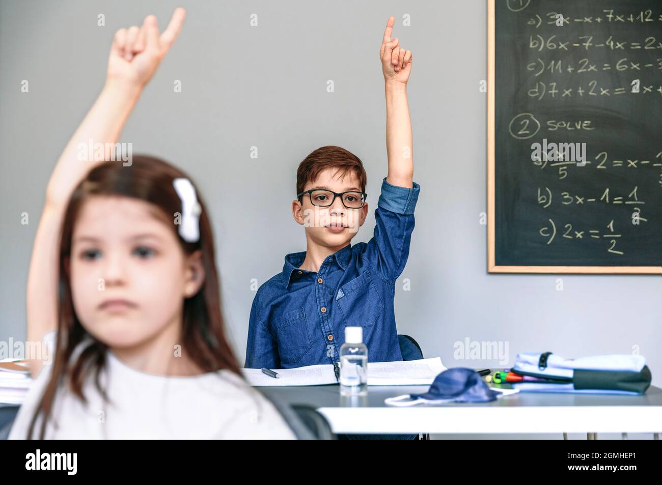 Students with mask on table raising hands at school Stock Photo