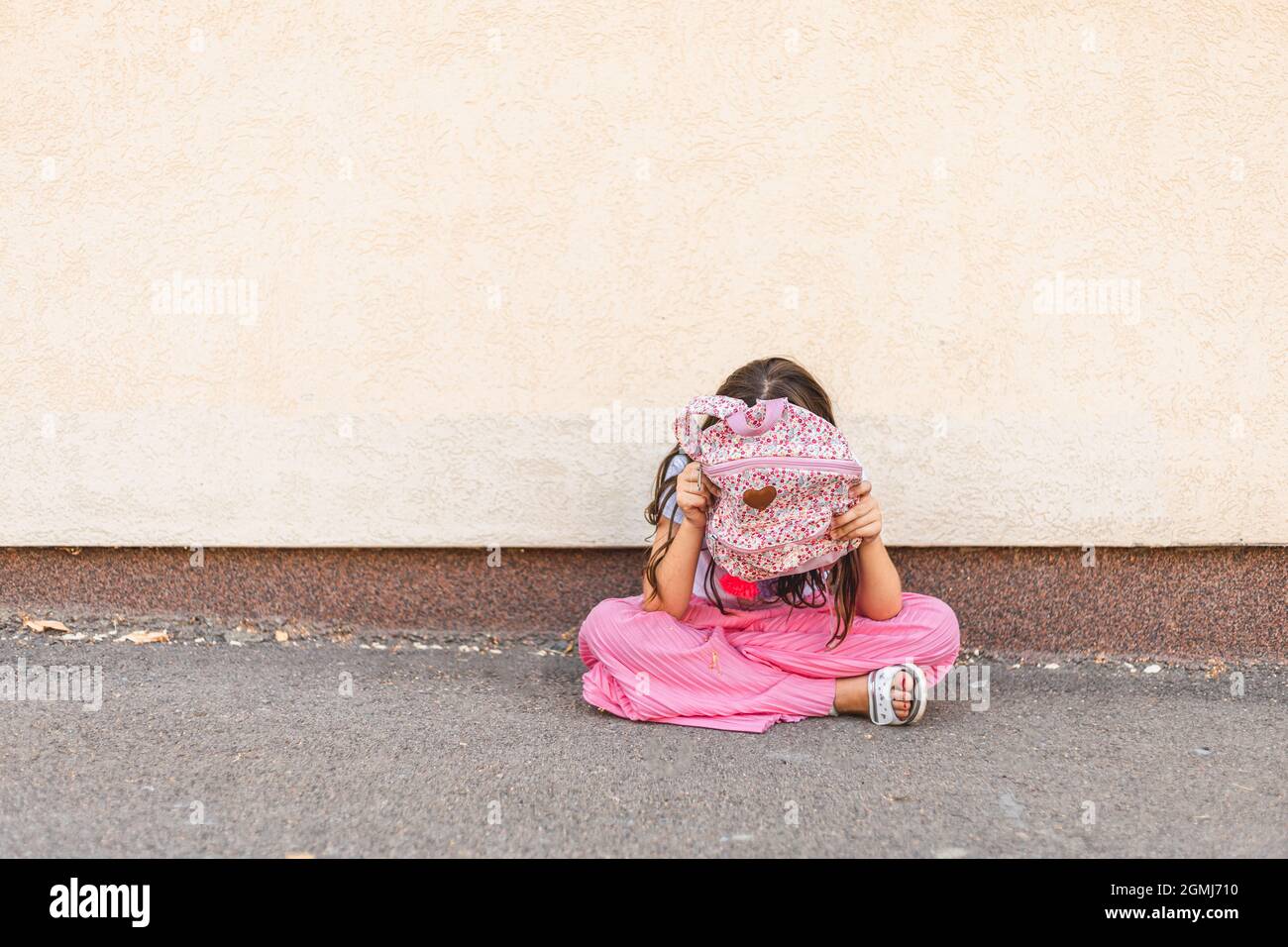 Little brunette girl hiding behind her rucksack, sitting alone and feeling sad Stock Photo