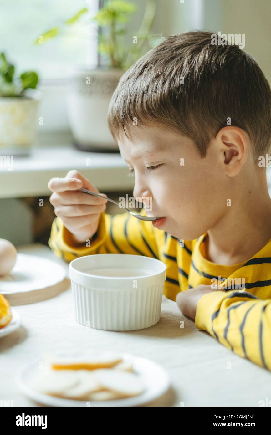 boy eats breakfast in the morning with cereals with milk. Morning breakfast in the kitchen before school. The boy is eating at the table by the window Stock Photo