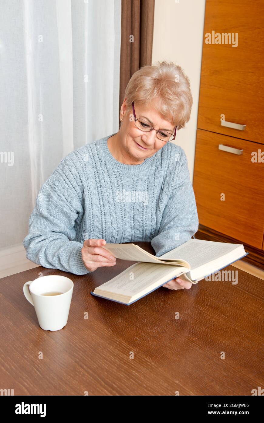 An elderly woman reading a book and drinking coffee at home Stock Photo
