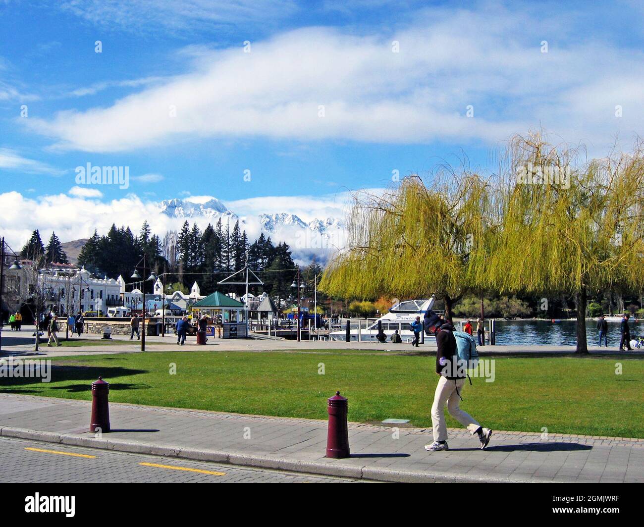 People walk along the waterfront in Queenstown, New Zealand during the spring with the trees starting to bloom and the mountains still covered in snow. Stock Photo