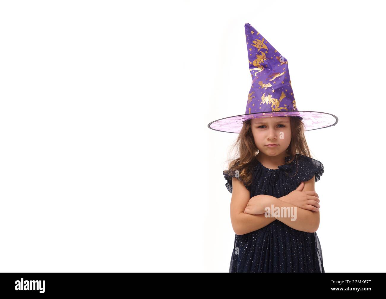 Portrait of winsome little witch angry upset girl wearing a wizard hat and dressed in stylish carnival dress, looking at camera posing with crossed ar Stock Photo
