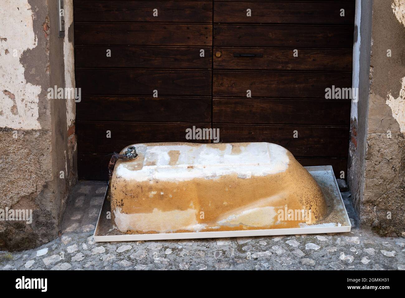 Old metal bath lyes upside down on cobblestone pavement outdoors in front of big wooden gate