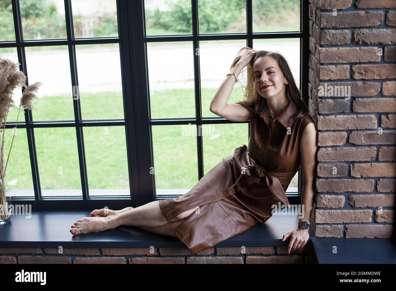 Beautiful young woman in brown leather dress sits on the windowsill ...