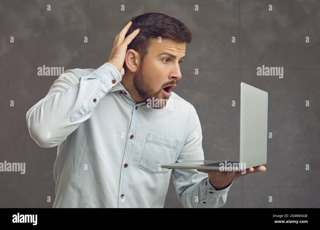 Shocked young man looking at screen of laptop computer that he's holding in hand Stock Photo