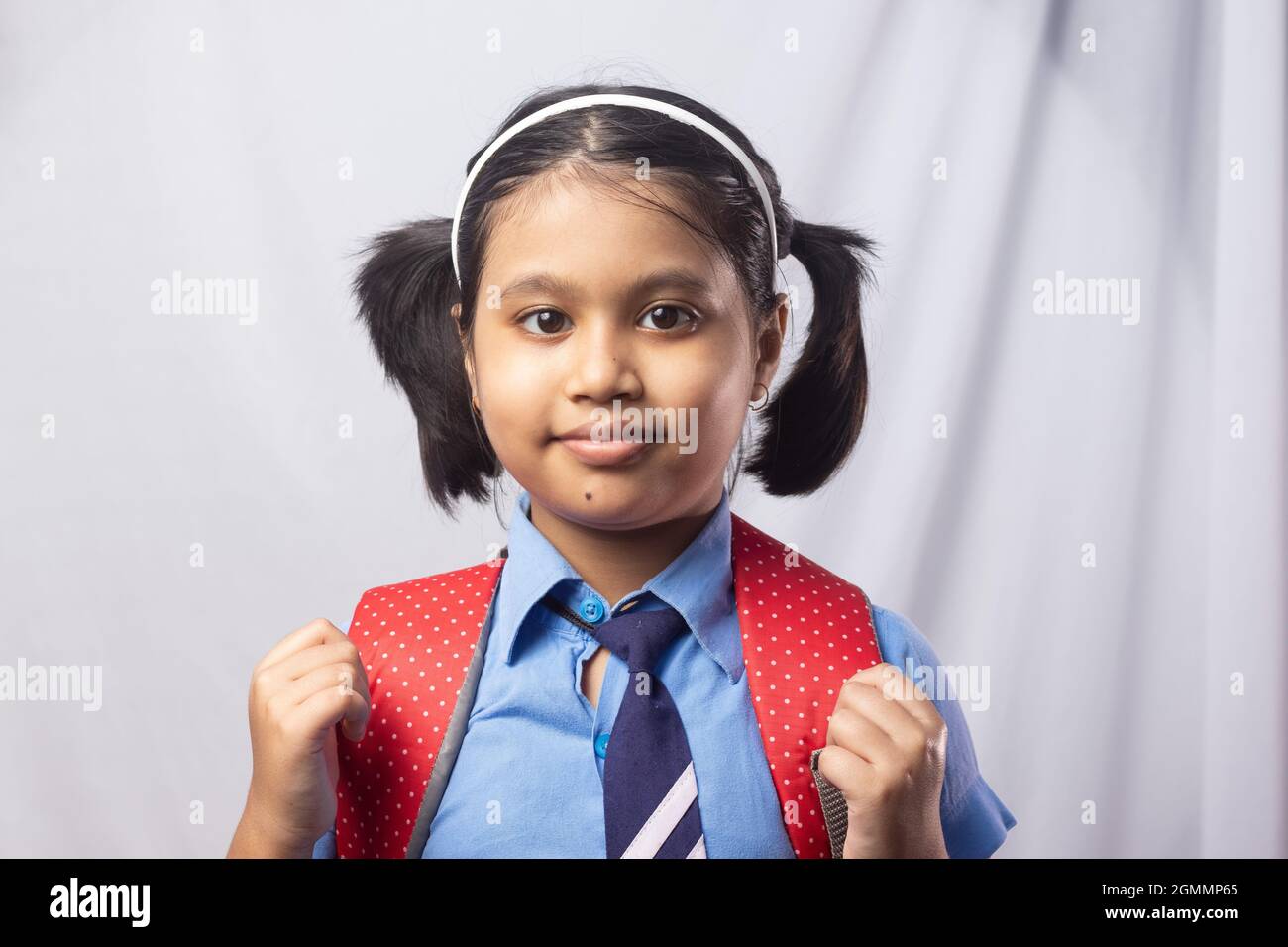 Portrait of a happy smiling Indian girl child student in blue school uniform with red bag on grey background Stock Photo
